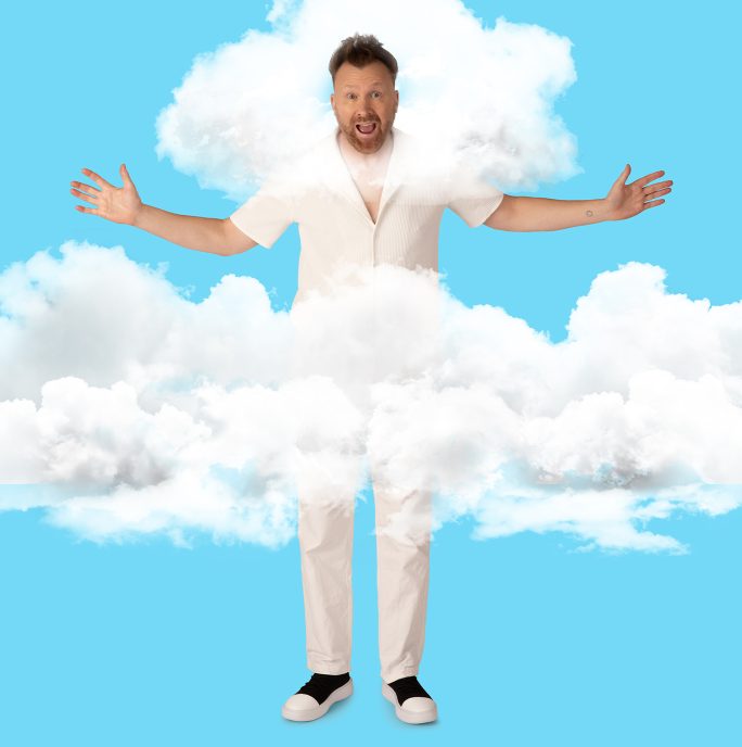 Jason Byrne, a white man dressed in white trousers and a white tshirt is standing against a bright blue backround surrounded by clouds