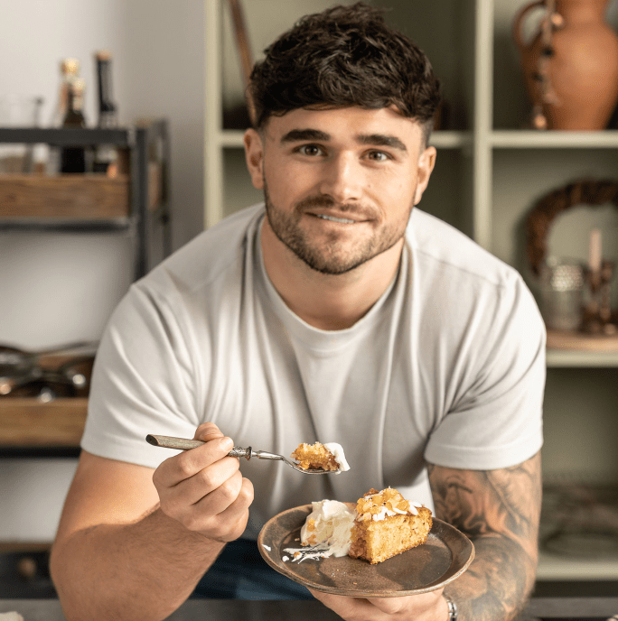 Matty Edgell standin in a kitchen leaning over a table holding a plate with a slice of cake